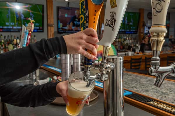 Bartender pouring draft beer from a branded tap handle into a pint glass at a sports bar, with multiple flat-screen TVs showing games in the background — a lively atmosphere for beer lovers.