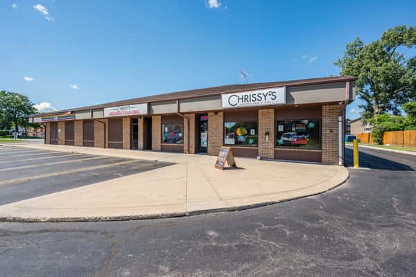 Exterior of Chrissy's Breakfast & Grill located in a strip mall, featuring a brick facade, large windows with neon signs, and a sandwich board welcoming customers.