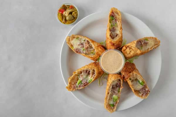 Golden-fried Philly cheesesteak egg rolls arranged around a creamy dipping sauce, filled with shaved steak, cheese, onions, and peppers — served with a side of pickled jalapeños.