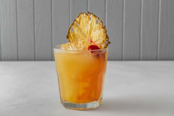 Refreshing orange cocktail served over ice in a short glass, garnished with a dried pineapple slice and cherry — a vibrant tropical drink perfect for summer sipping.