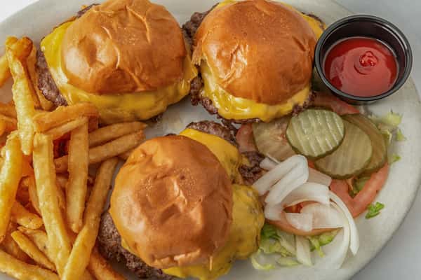 Three juicy cheeseburger sliders with melted cheddar on toasted buns, served with seasoned fries, pickles, onions, tomatoes, and a side of ketchup — a hearty American classic.