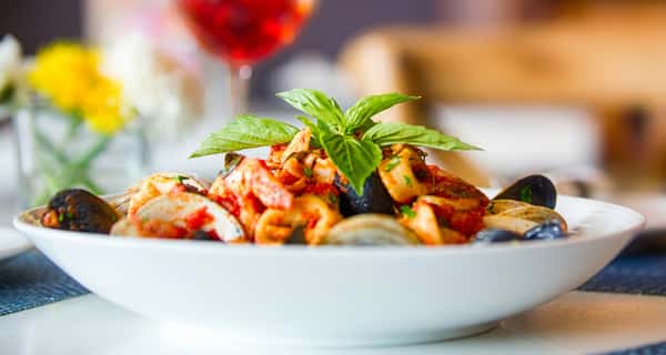 Seafood pasta with mussels, clams, squid, red sauce, and basil, in a white bowl with red wine in the background.