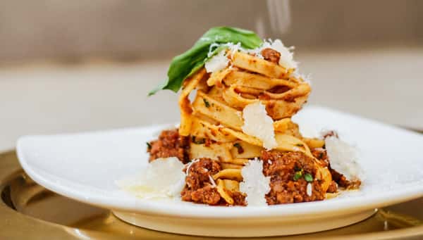 Tagliatelle pasta with Bolognese sauce, Parmesan shavings, and a fresh basil leaf on a white plate.