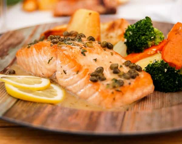 Salmon fillet with capers, lemon slices, roasted potatoes, broccoli, and carrots on a wooden plate.