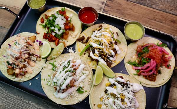 A vibrant tray of assorted tacos featuring various toppings such as cilantro, onions, cheese, crema, pickled onions, and limes, served with salsa and guacamole on the side.