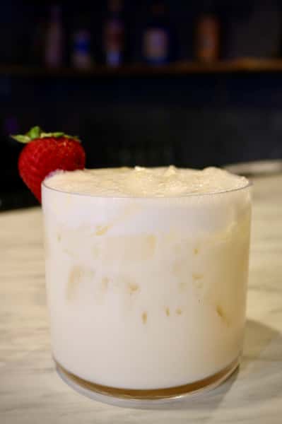 A creamy white cocktail served over ice in a short glass, garnished with a fresh strawberry on the rim.