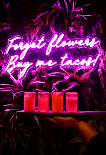 Four colorful cocktails with chili-salt rims held on a tray beneath a neon pink sign reading "Forget flowers, buy me tacos!"