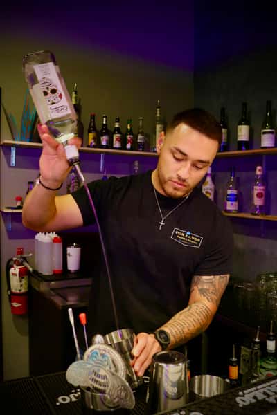 Bartender pouring liquor into a shaker behind the bar, surrounded by bottles and bar tools under warm lighting.