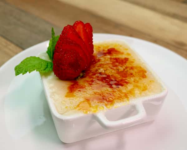 A beautifully caramelized crème brûlée served in a small white dish, garnished with a sliced strawberry and a fresh mint sprig.