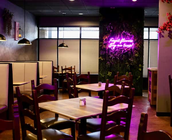 Cozy restaurant interior with wooden tables, booth seating, and a neon sign reading "Forget flowers, buy me tacos" on a plant-covered wall.