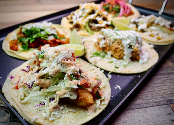 A vibrant tray of assorted tacos featuring various toppings such as cilantro, onions, cheese, crema, pickled onions, and limes, served with salsa and guacamole on the side.