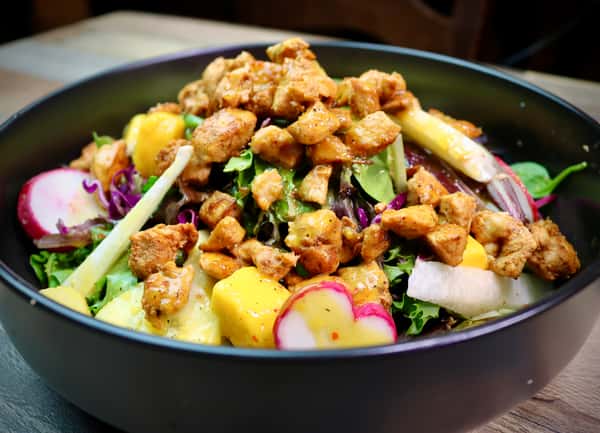 A fresh and colorful salad topped with grilled chicken pieces, featuring mixed greens, radishes, mango chunks, and other vibrant vegetables.
