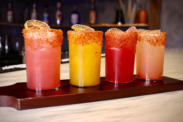 Four colorful cocktails in tall glasses rimmed with chili powder and garnished with dried lime slices, displayed on a wooden tray.