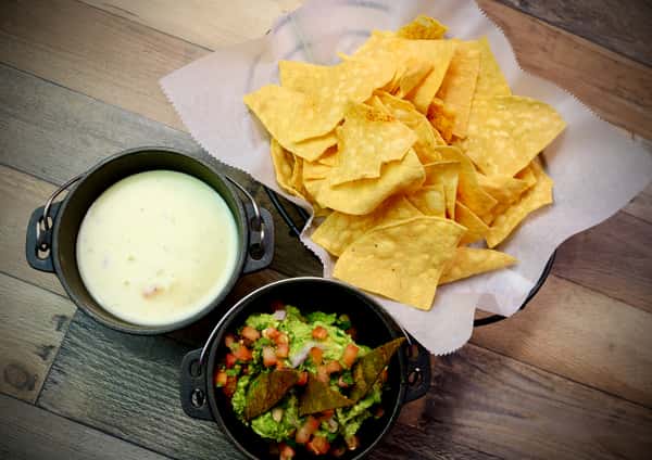A serving of crispy tortilla chips paired with creamy queso dip and fresh guacamole topped with diced tomatoes and tortilla triangles, presented in small black bowls.
