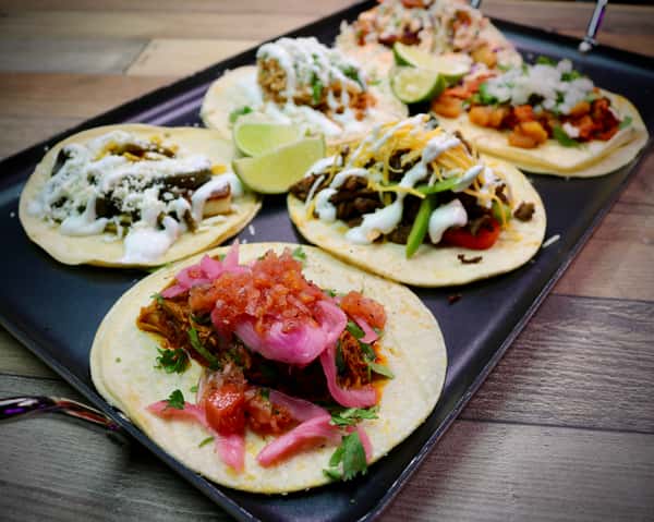A vibrant tray of assorted tacos featuring various toppings such as cilantro, onions, cheese, crema, pickled onions, and limes, served with salsa and guacamole on the side.