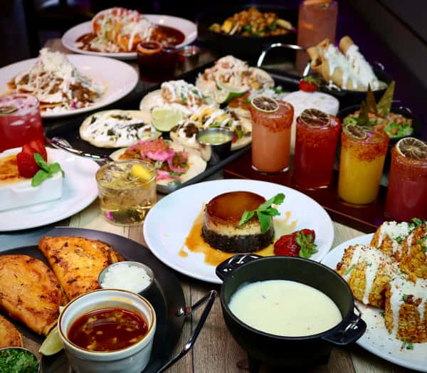 Colorful Mexican feast with tacos, birria, elote, queso dip, flautas, desserts, and vibrant cocktails arranged on a wooden table.