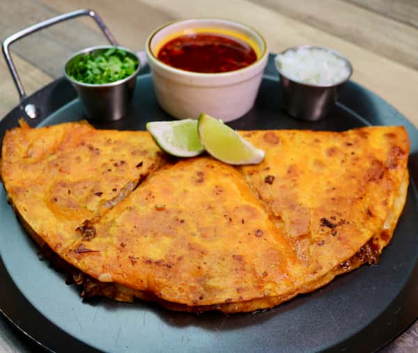Crispy birria tacos served with a rich red consommé, fresh cilantro, lime wedges, and onions on a dark plate.