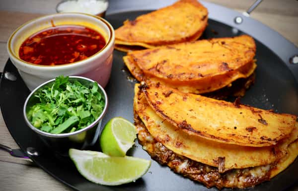 Crispy birria tacos served with a rich red consommé, fresh cilantro, lime wedges, and onions on a dark plate.