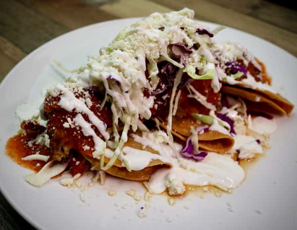 Crispy flautas topped with shredded cabbage, crema, cotija cheese, and red salsa on a white plate.