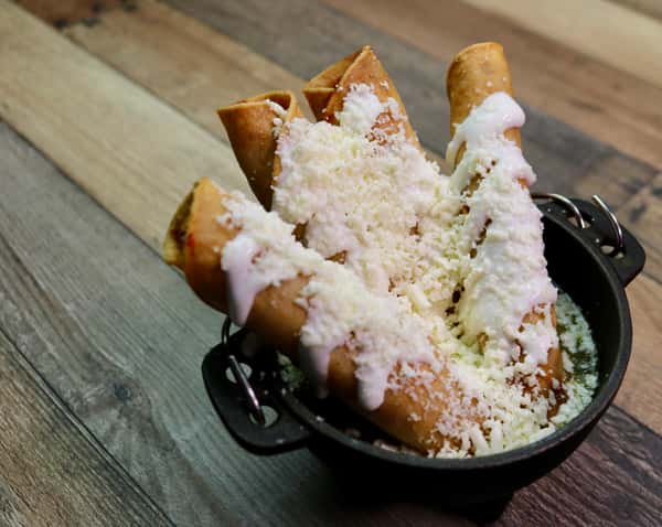 Golden crispy flautas topped with crema and crumbled cotija cheese, served in a small black bowl.