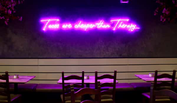 A cozy restaurant interior with a neon pink sign reading "Tacos are cheaper than Therapy" glowing above wooden tables and chairs.