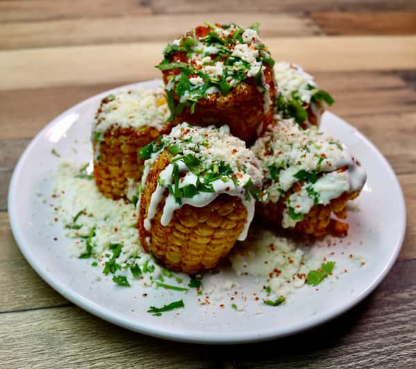 A plate of elote (Mexican street corn), served in small cob sections topped with creamy sauce, crumbled cheese, chili powder, and fresh cilantro.