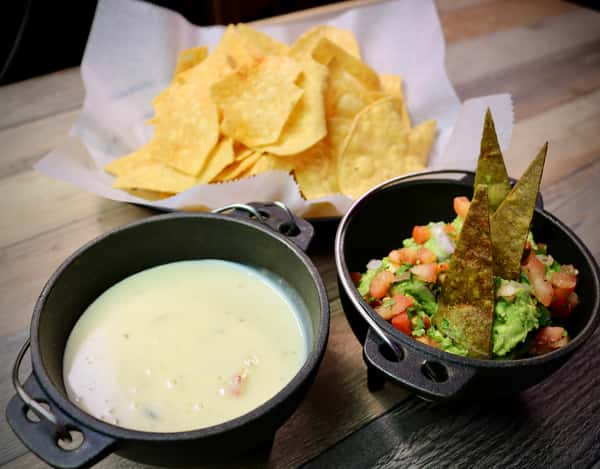 A serving of crispy tortilla chips paired with creamy queso dip and fresh guacamole topped with diced tomatoes and tortilla triangles, presented in small black bowls.