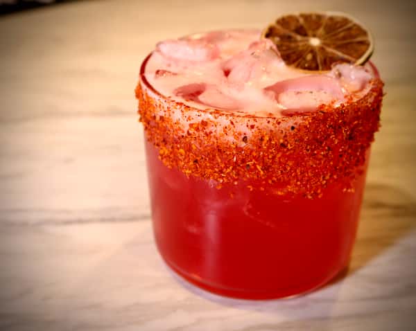 Vibrant red cocktail with a chili-lime rim, ice cubes, and a dried lime garnish served in a short glass on a marble surface.