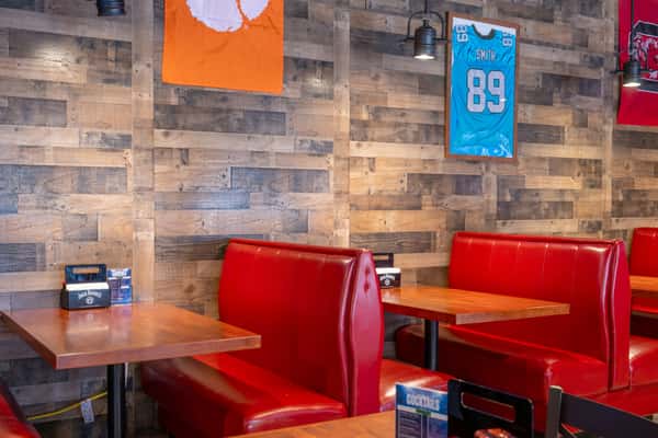 Bright dining area with red booth seating and wooden tables, wall-mounted TVs showing sports, hanging pendant lights, and large windows letting in natural light.