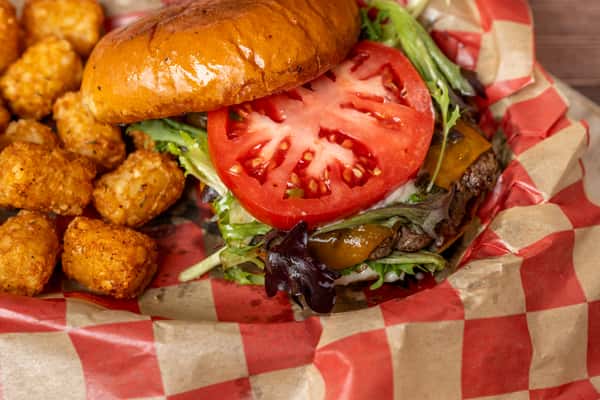 Cheeseburger with lettuce, tomato, and melted cheddar on brioche bun, served with crispy tater tots on checkered paper.