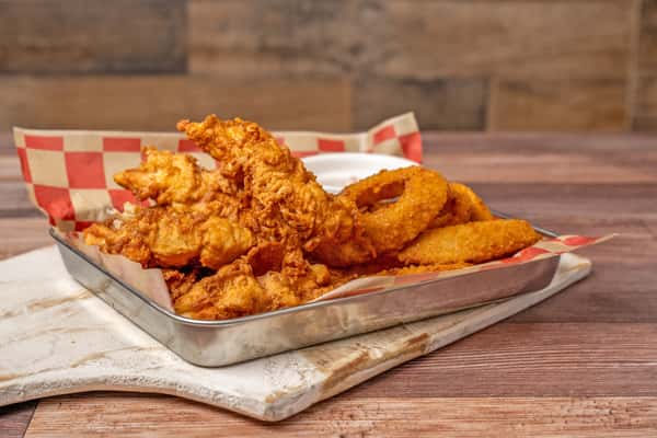 Metal tray filled with crispy fried chicken tenders and golden onion rings, lined with red-and-white checkered paper and served with a side of dipping sauce.
