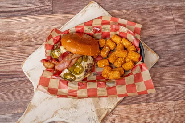 Bacon cheeseburger topped with jalapeños and creamy sauce, served with tater tots in a checkered basket.