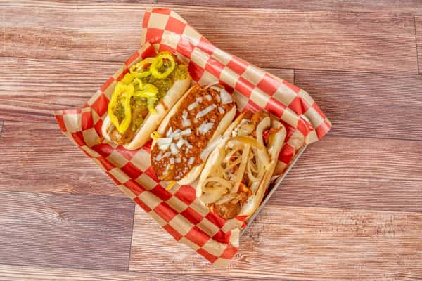 Three hot dogs in buns served in a metal tray with checkered paper, topped with chili and diced onions, pickles and peppers, and sautéed onions with bacon.