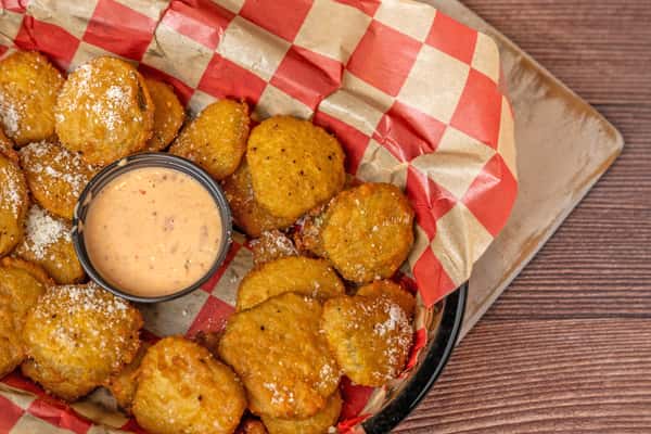 Top-down view of a basket of fried pickle chips dusted with grated cheese, served on red-and-white checkered paper with a side of creamy dipping sauce.