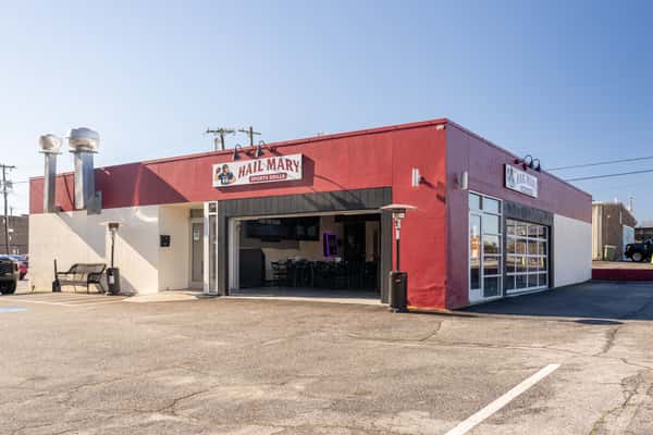 Exterior view of a red and white building labeled "Hail Mary Sports Grille," with large open garage-style doors, patio heaters outside, and a mostly empty parking lot under a clear sky.