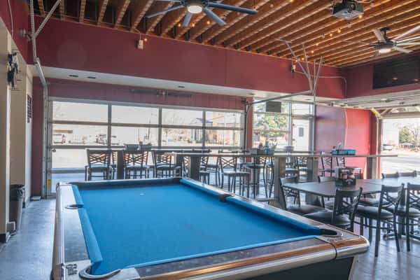 Interior of a casual bar with a blue felt pool table, high-top tables, large open garage doors, and ceiling fans.
