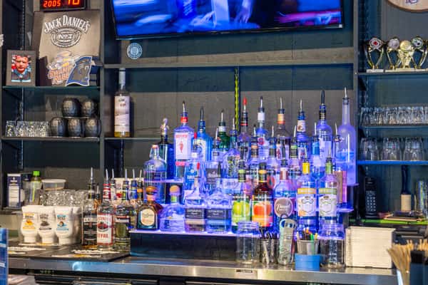 Back bar display with assorted liquor bottles arranged on illuminated shelves, glassware, and bar equipment beneath a mounted TV screen.
