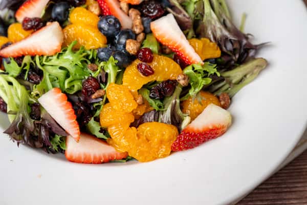 Mixed greens salad with strawberries, blueberries, mandarin oranges, dried cranberries, and candied pecans on white plate.