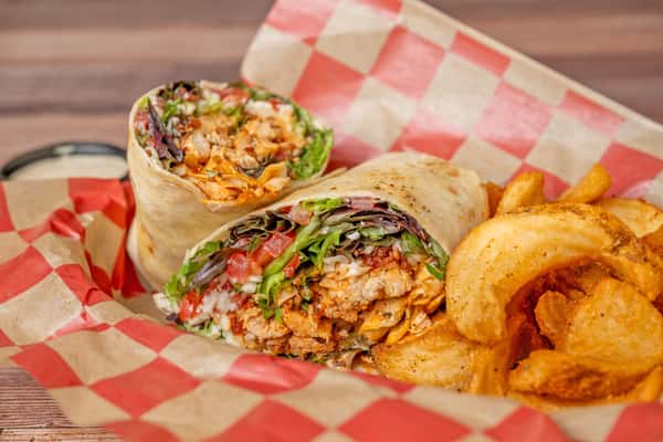 Close-up of a sliced chicken wrap filled with seasoned grilled chicken, lettuce, tomato, and shredded cheese, served in a basket lined with red-and-white checkered paper alongside golden potato wedges and a side of dipping sauce.