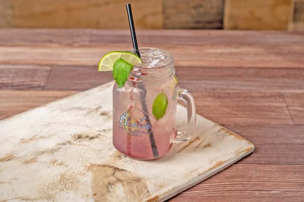 Pink cocktail in a mason jar with ice, garnished with a lime wedge and fresh mint, on a wooden board.