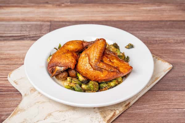 Roasted chicken halves served over crispy Brussels sprouts on a white plate atop a wooden table.