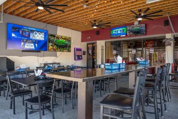 Spacious bar and dining area with high-top tables and chairs, wall-mounted TVs showing sports, exposed wood-beam ceiling with large ceiling fans, and baskets of utensils on the tables.