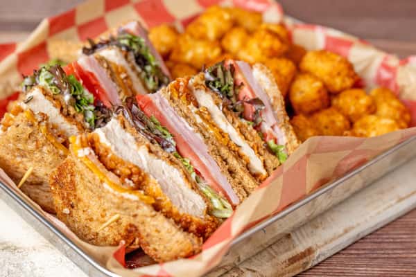 Close-up of a sliced club-style sandwich stacked with crispy fried chicken, ham, cheese, lettuce, and tomato on toasted bread, served in a metal tray with red-and-white checkered paper and a side of tater tots.