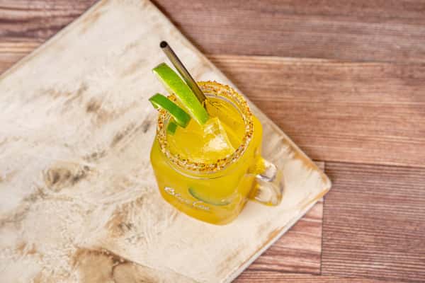 Mango margarita in a mason jar with chili salt rim, lime wedges, ice, and a metal straw on a wooden table.