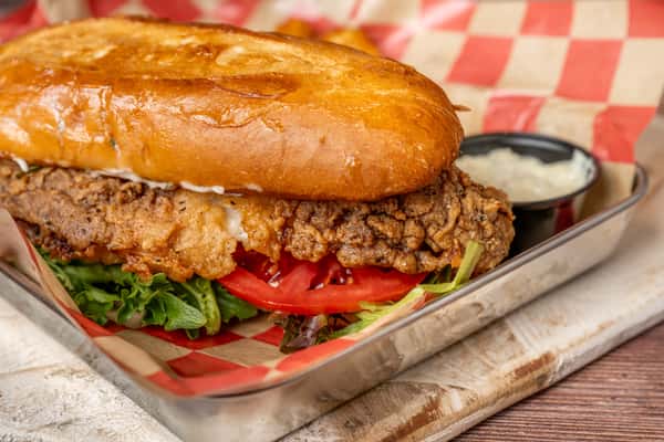 Close-up of a crispy fried chicken sandwich on a toasted bun with lettuce and tomato, served in a metal tray with checkered paper and a side of dipping sauce.
