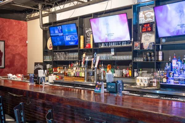 Wide view of a bar interior with a long wooden counter, shelves stocked with liquor bottles and glassware, multiple beer taps, and several wall-mounted TVs above the back bar.