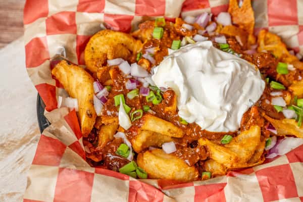 Chili cheese fries topped with sour cream, diced red onions, and green onions in a checkered basket.