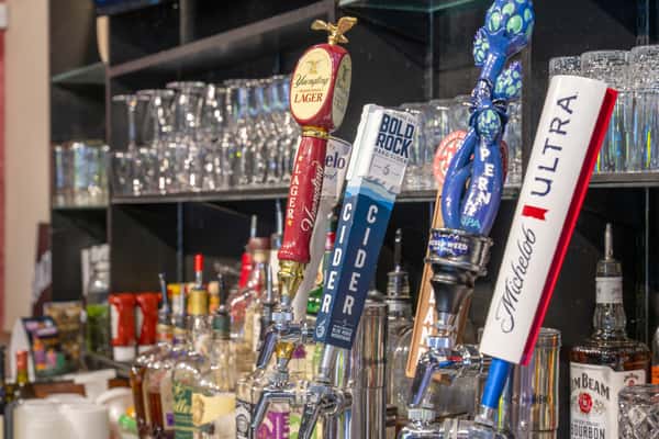Close-up of several beer taps at a bar, including handles for lager, cider, IPA, and Michelob Ultra, with liquor bottles and glassware in the background.