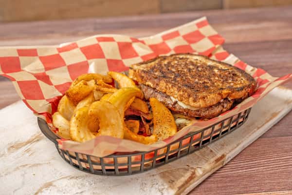 Grilled patty melt sandwich with melted cheese, served with seasoned fries and onion rings in a checkered basket.