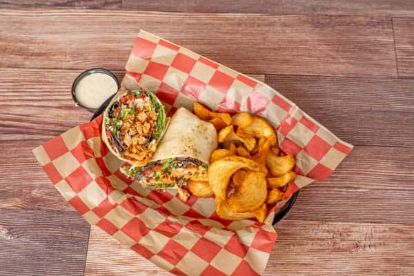 Close-up of a sliced chicken wrap filled with seasoned grilled chicken, lettuce, tomato, and shredded cheese, served in a basket lined with red-and-white checkered paper alongside golden potato wedges and a side of dipping sauce.